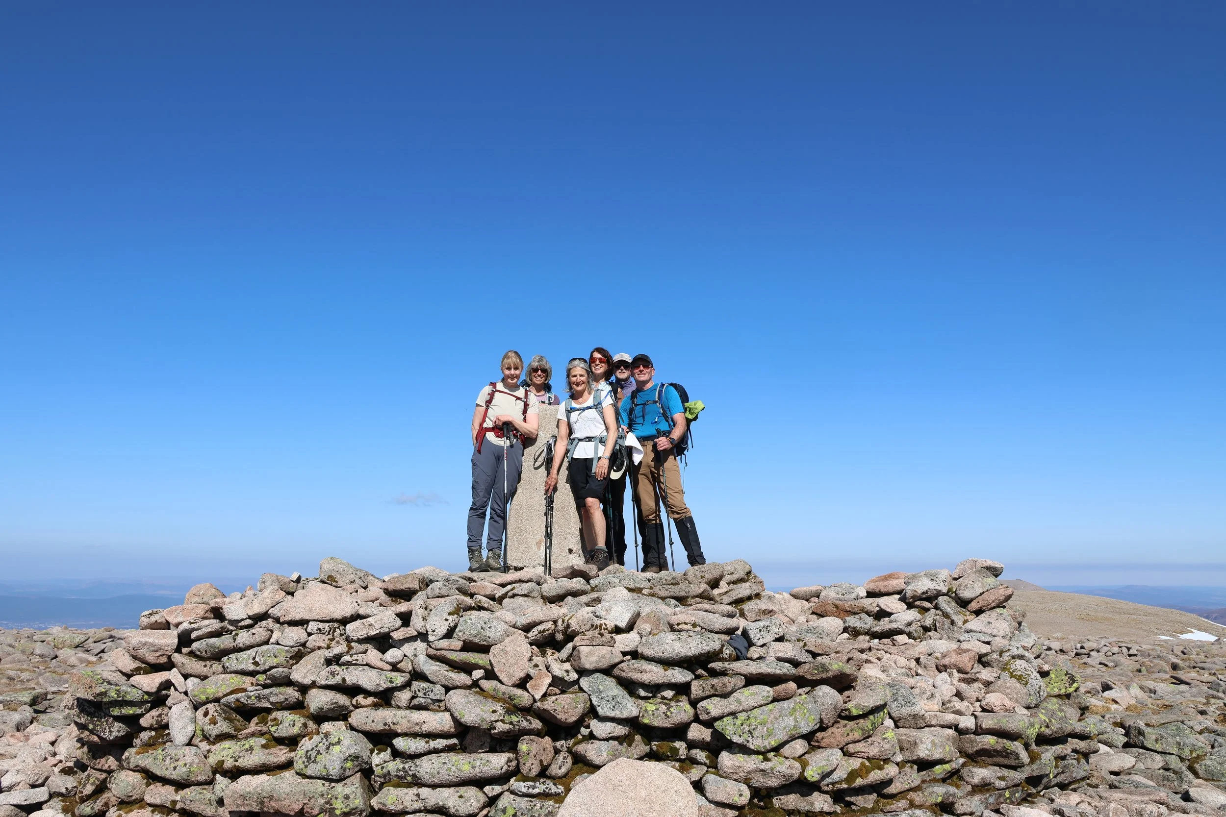 A group of six hikers stands on a rocky summit, with a blue sky above and distant landscape below.
