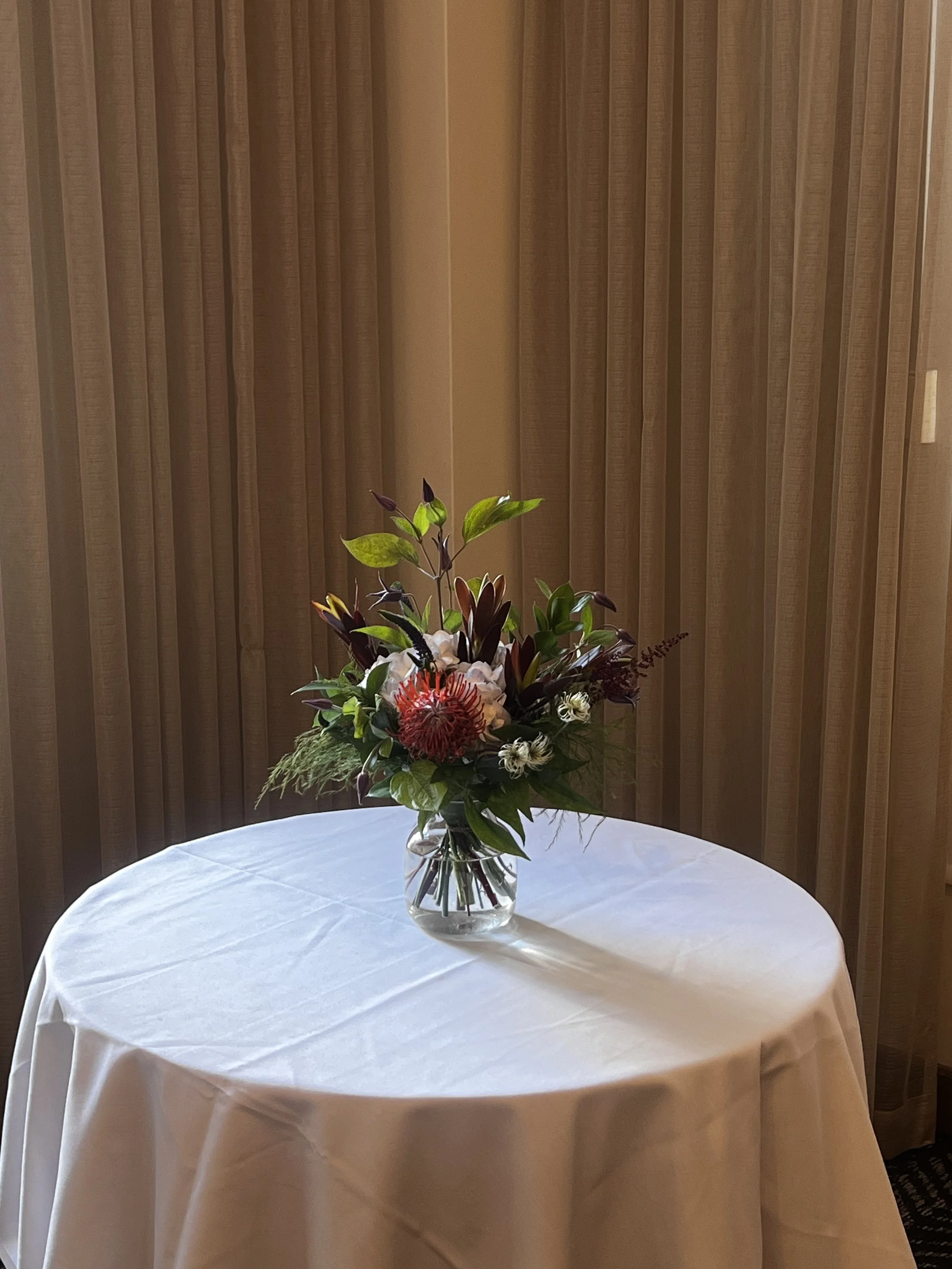 Composition florale sur mesure designé pour un événement corporatif au club st-james au centre-ville de montéal. Vase de fleurs sur une table ronde avec nappe blanche devant des rideaux beige.