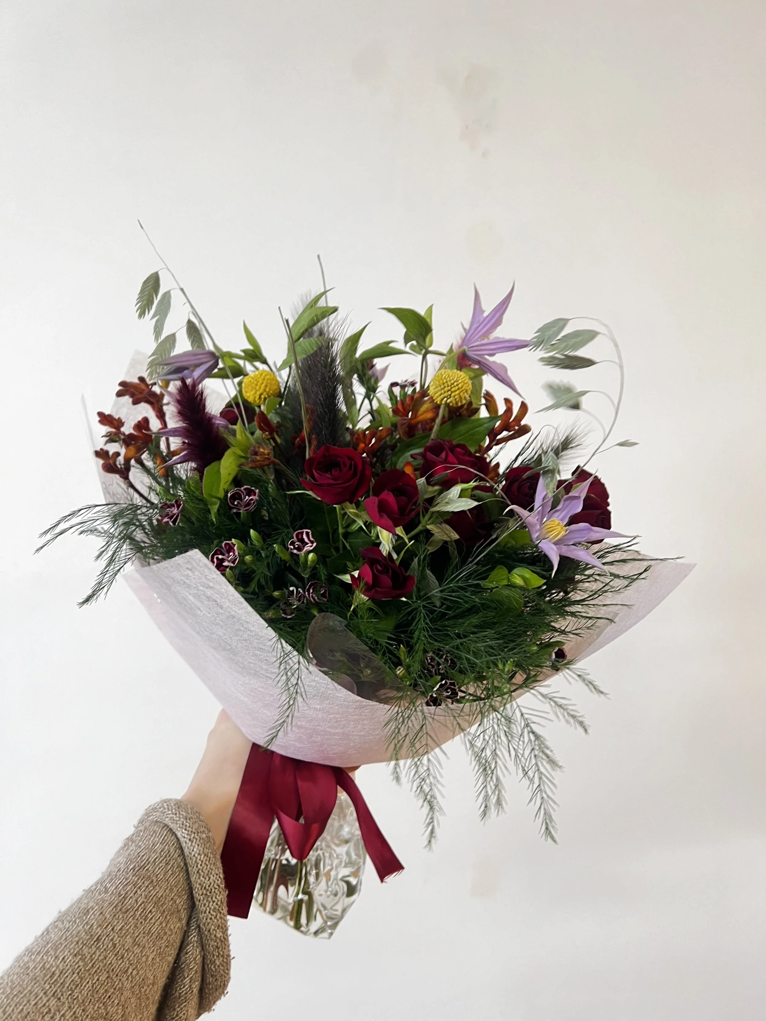 Un bouquet de fleurs avec des roses rouges, des fleurs violettes et des plantes vertes, enveloppé dans du papier avec un ruban rouge.