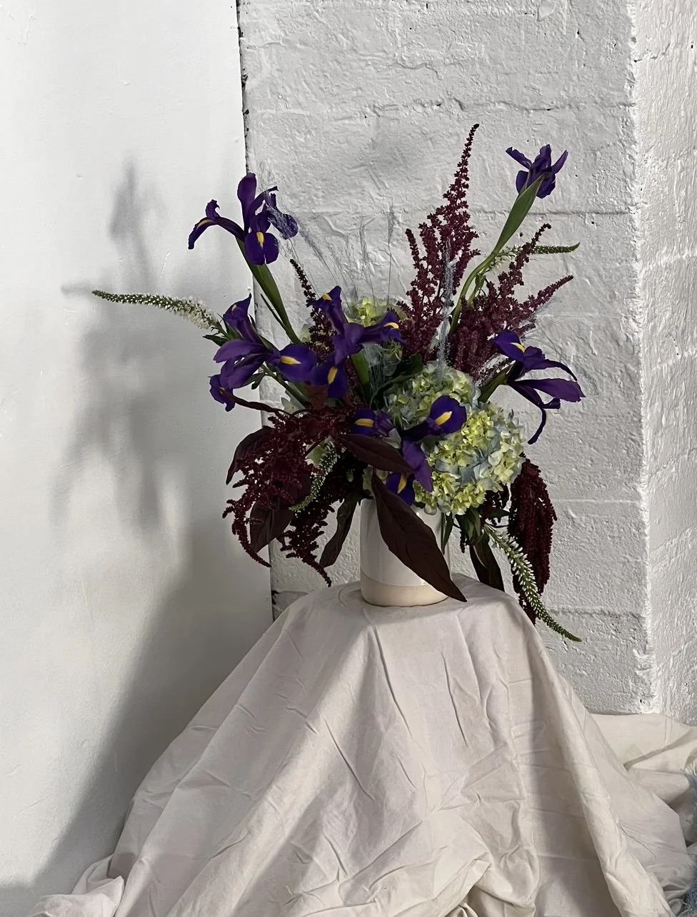 Composition florale avec iris, hortensias et feuillage dans un vase blanc, placé sur une table avec une nappe beige, contre un mur en briques blanches.
