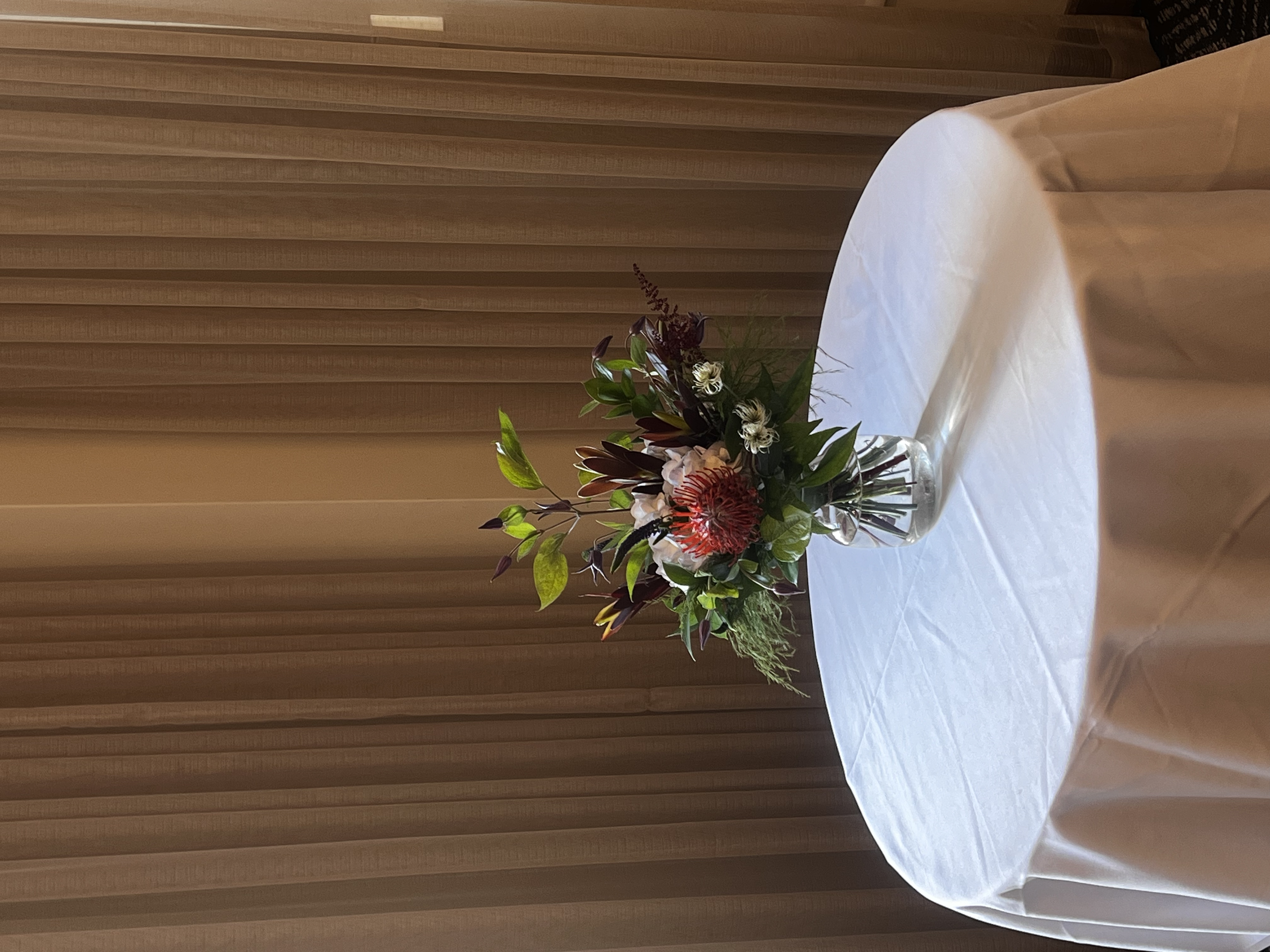 Un arrangement floral dans un vase en verre sur une table arrondie avec une nappe blanche, en intérieur, avec des rideaux en tissu brun à l'arrière-plan, pour un evenement.