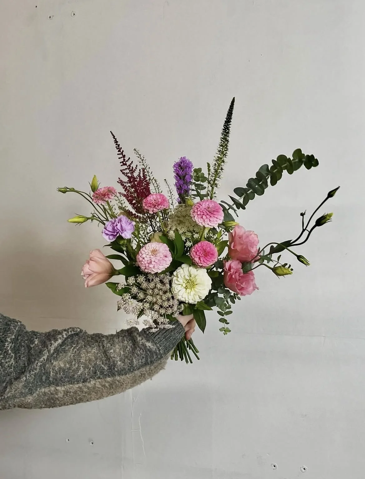 Main dans une manche de pull gris clair, tenant un bouquet de fleurs composé de roses, dahlias, lavande, et feuillage vert, contre un mur blanc.