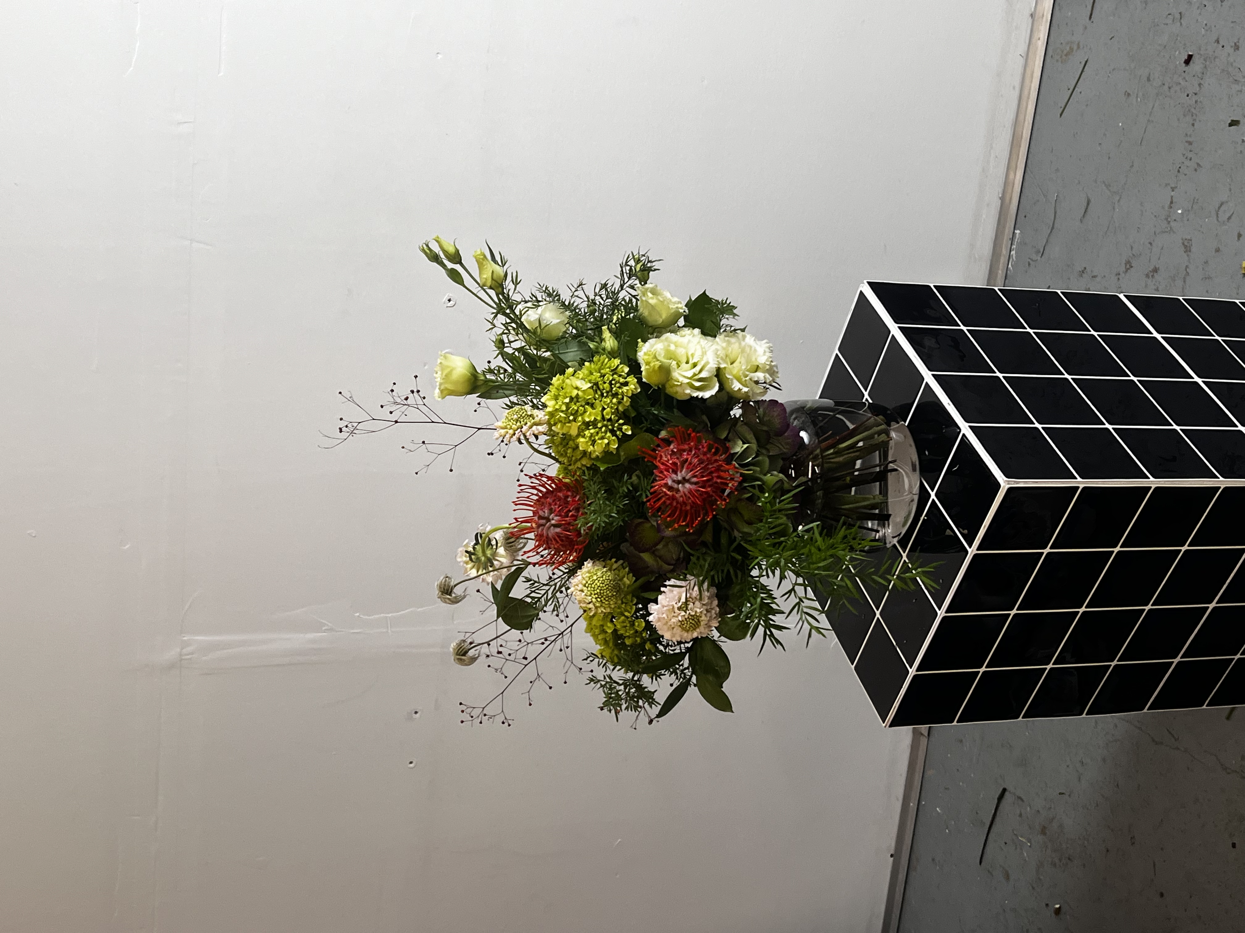 Bouquet de fleurs variées dans un vase transparent, placé sur une table noire à carreaux blancs, devant un mur blanc.