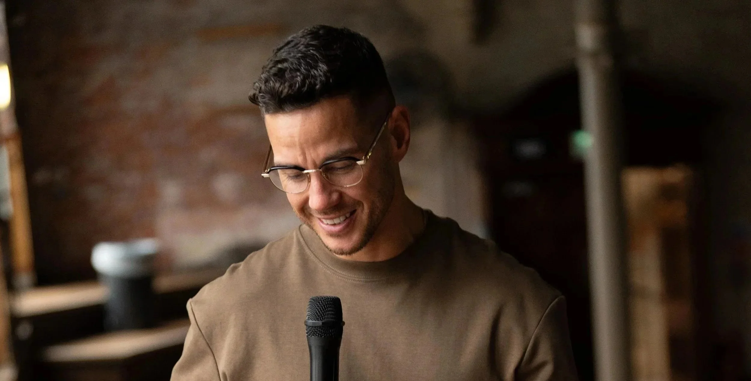 Scott Thomas wearing glasses looking down at a microphone in his hand inside a cosy, dimly-lit room with a brick wall background.