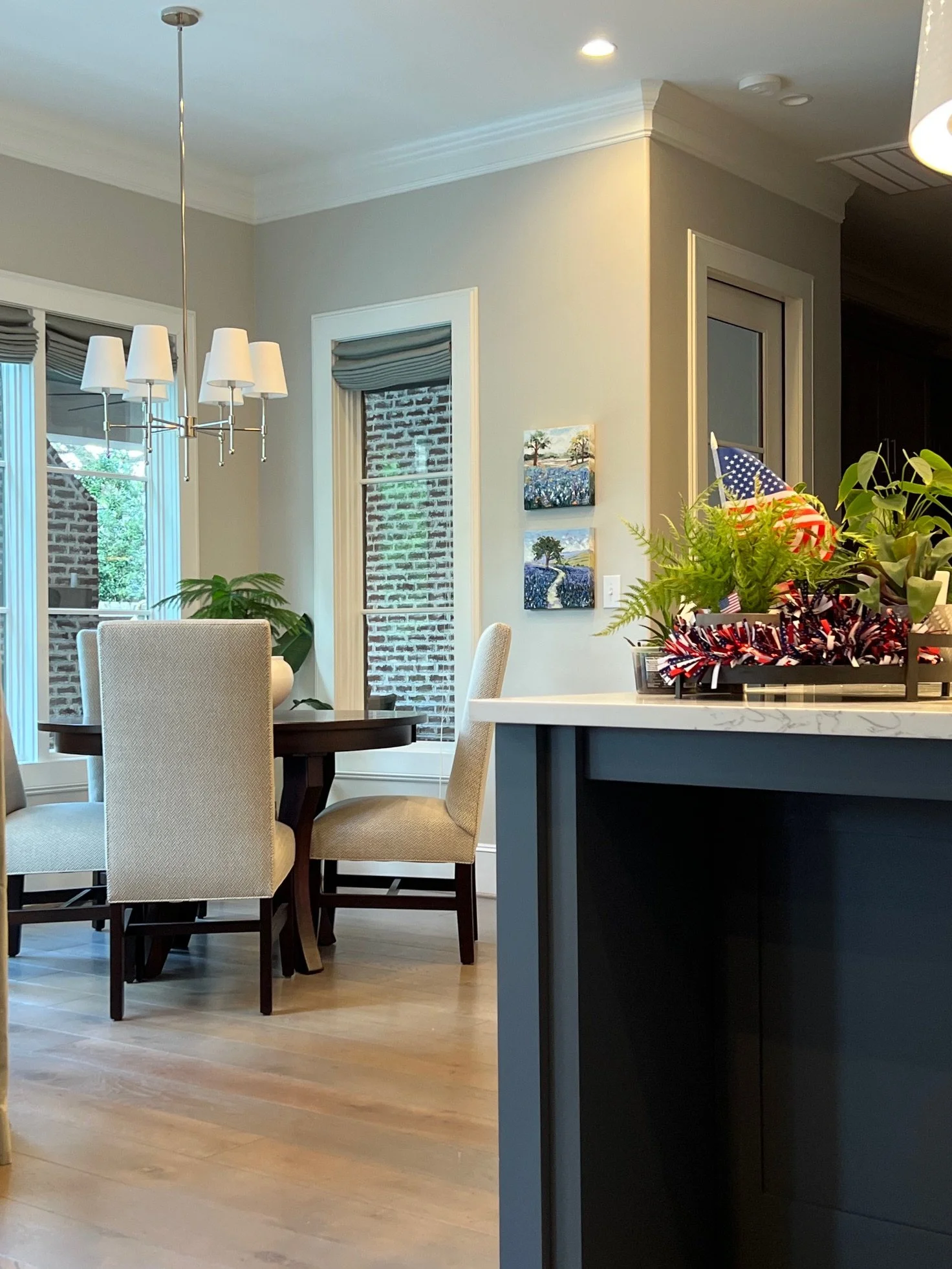 A dining area with a round table and four upholstered chairs, a chandelier hanging from the ceiling, large windows with Roman shades, wall art, and a kitchen counter with a potted plant and patriotic decorations.
