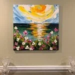 Painting of a sunset over water with flowers in the foreground hanging on a beige wall.