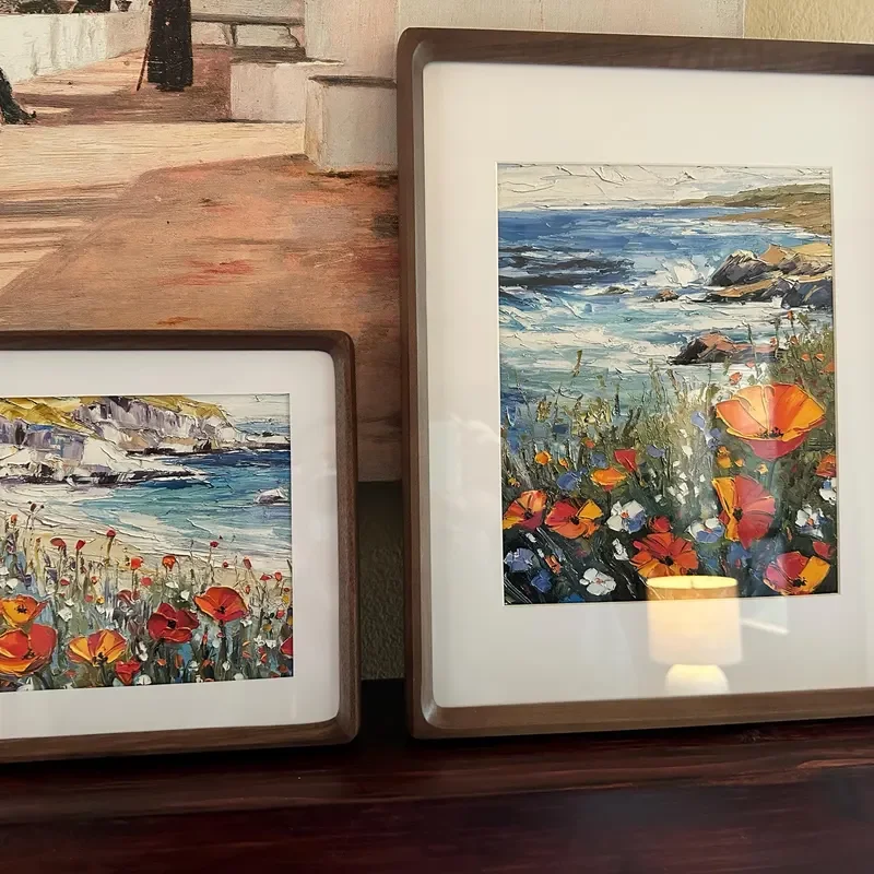 Two framed landscape paintings of coastal scenes with flowers in the foreground, one showing a rocky shoreline with waves and the other depicting a beach with flowers and cliffs.
