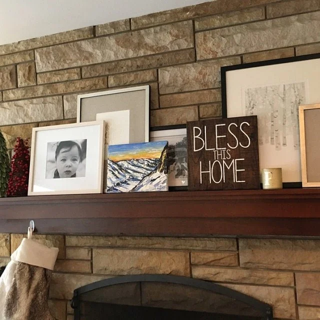 A wooden mantelpiece decorated with framed photographs, artwork, and a small sign that reads "Bless This Home," set against a stone brick fireplace wall.