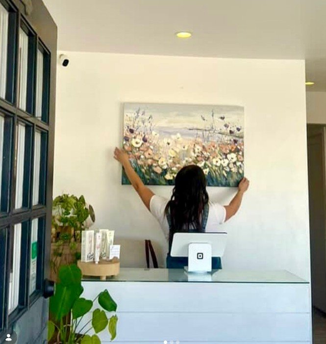 A woman hanging a large floral painting on a white wall in an interior space, with plants and a white desk in the foreground.