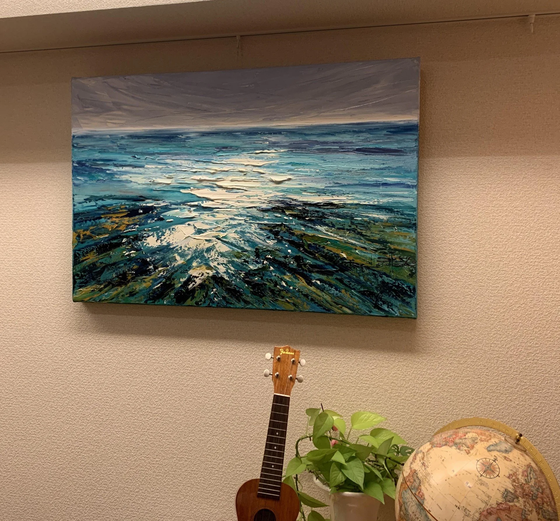 Wall-mounted painting of a seascape with stormy clouds and ocean waves, a ukulele hanging in front, and a potted plant on a table with a globe, located in a room with beige walls.