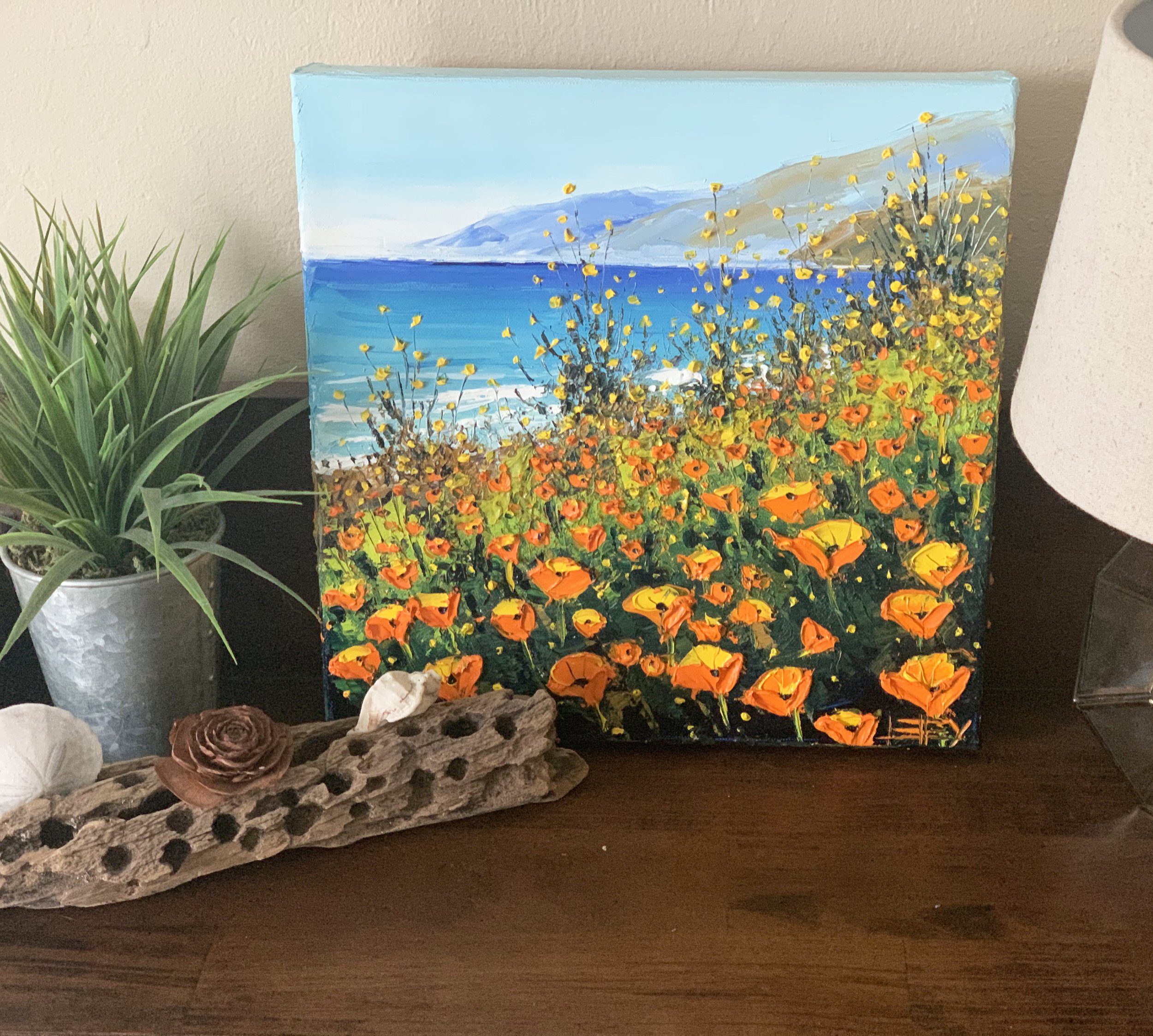 A landscape painting on canvas depicts a beach scene with mountains in the distance, blue sky, and yellow flowers in the foreground, positioned on a dark wooden surface.