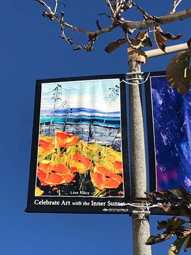 A street banner displaying an artwork titled "Celebrate Art with the Inner Sunset" by Lisa Elley, featuring a colorful landscape of orange flowers, a blue sky, and distant mountains, hanging from a street pole against a clear blue sky.
