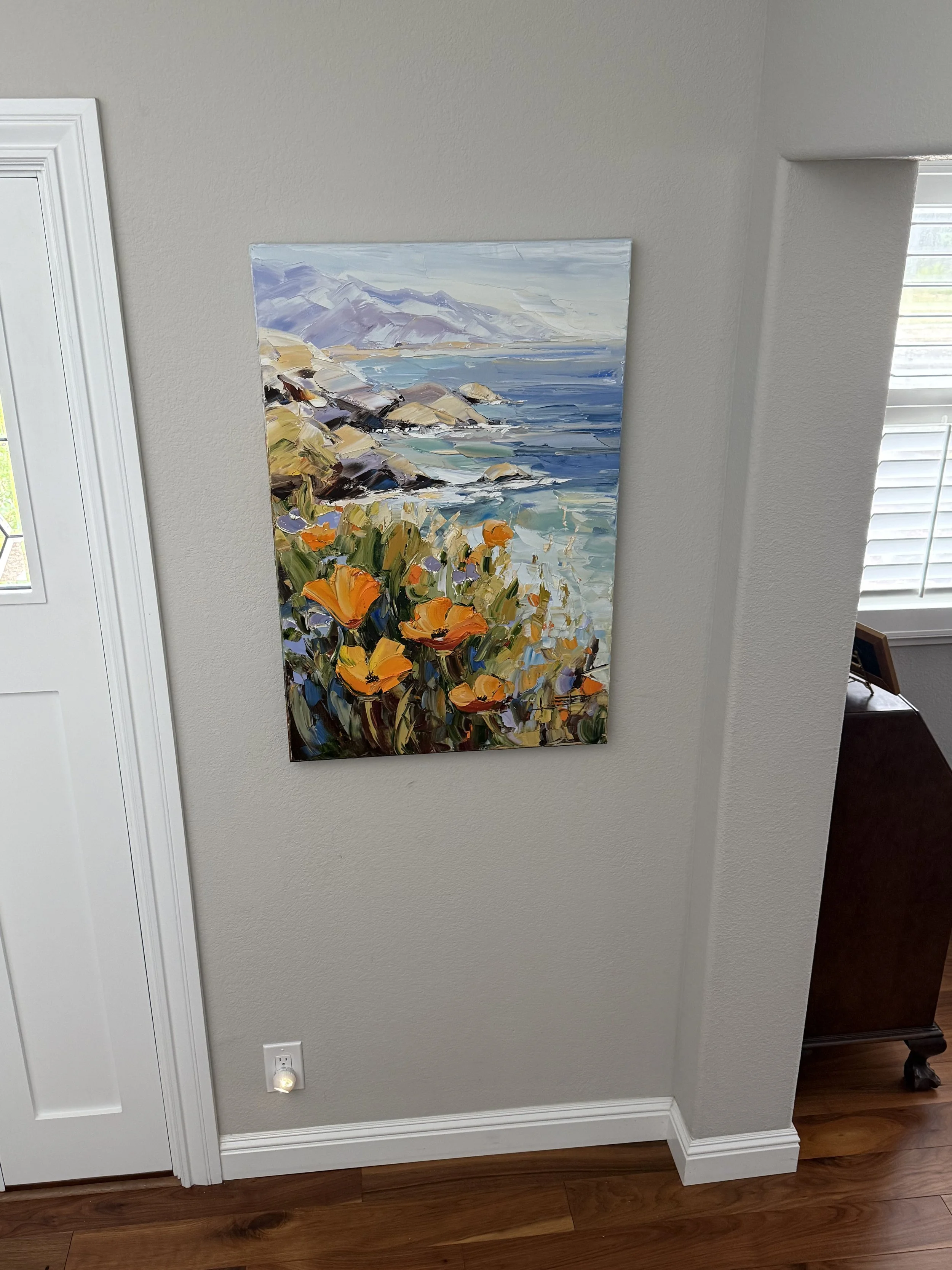 A colorful landscape painting of a mountain, ocean, and flowers on a white wall near a door and window in a room with wooden flooring.