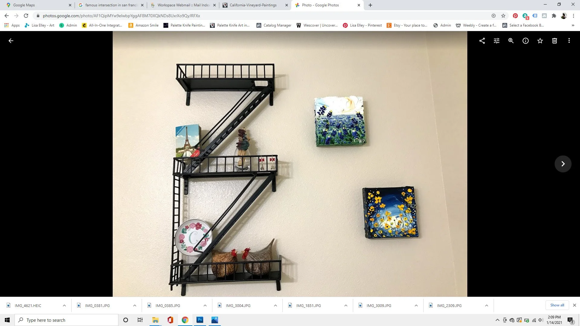 Decorative wall display with a black metal wall shelf shaped like the letter 'Z', holding small decorative items like miniatures and a plate, and surrounded by framed paintings of flowers, a Paris cityscape, and a nighttime floral scene on a beige wa