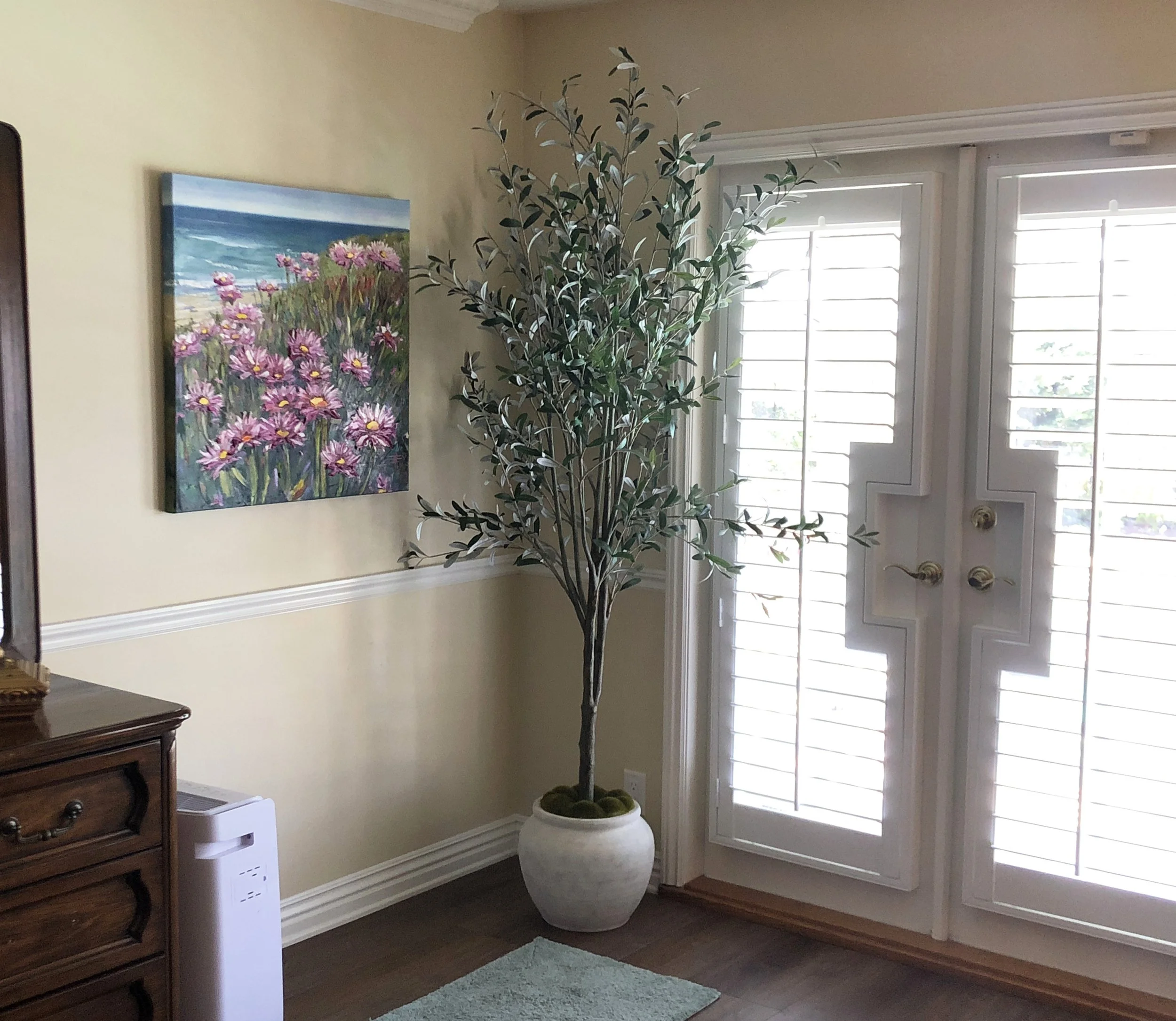 A tall potted plant with green leaves in a white pot stands next to a window with white shutters, a colorful painting of pink flowers and ocean scene hangs on the beige wall, and a piece of wooden furniture is partially visible on the left side of th