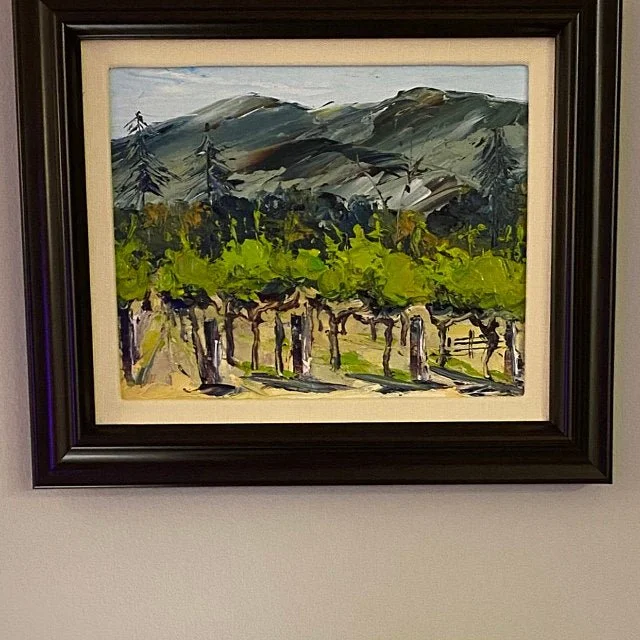 A framed watercolor painting of a rural landscape featuring rows of grapevines, green trees, and mountains in the background.