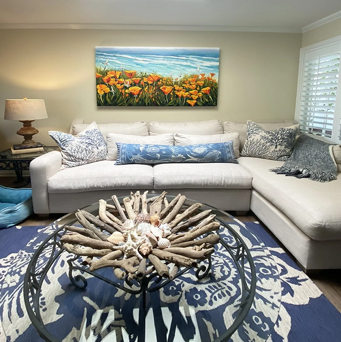 Living room with white sectional sofa, decorative pillows, a round glass-top table with driftwood and seashells, blue patterned rug, painting of a flower field on the wall, and windows with white blinds.
