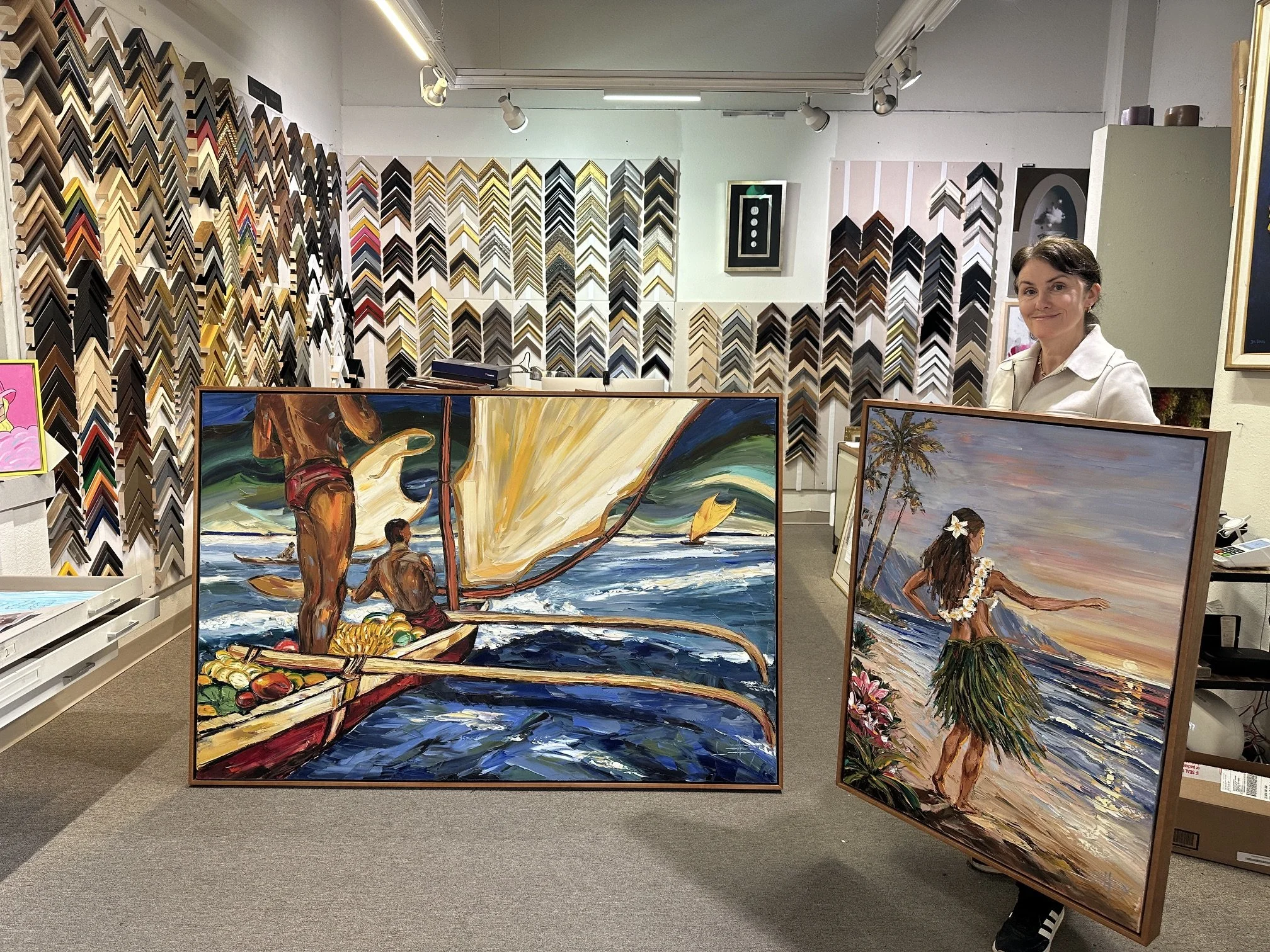 Interior of an art gallery with framed paintings and sample color frames on the wall. There are three standing paintings in the foreground, showing tropical scenes, people on the beach with sailboats, and a woman with flowers. A woman with short dark hair and a white shirt is standing and smiling.