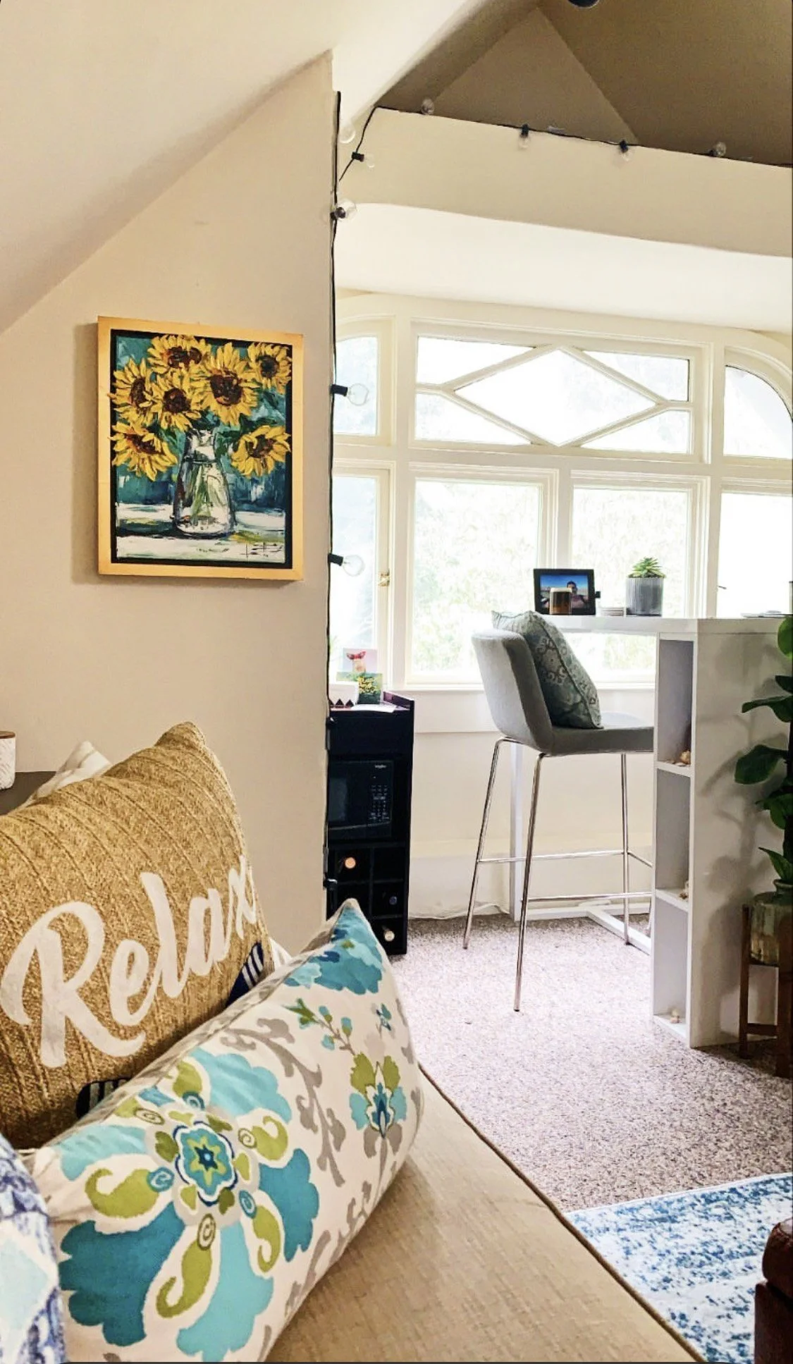 Interior view of a cozy apartment with a wall art piece of sunflowers, a window seat with a small table, potted plant, and a chair, and a cushioned area with pillows, including one with the word 'Relax'.
