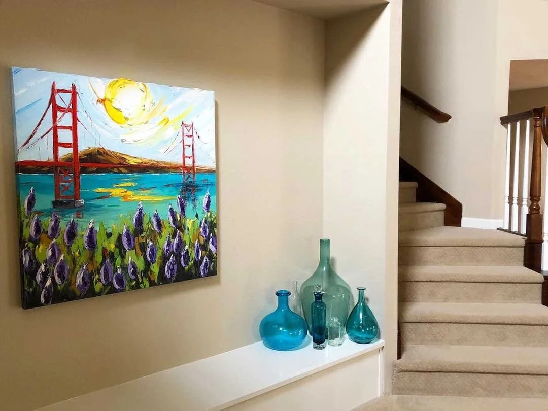Colorful painting of the Golden Gate Bridge with purple flowers in the foreground, mounted on a beige wall in a home interior. Three decorative glass vases in shades of blue and green sit on a white shelf below the painting, next to a staircase with 
