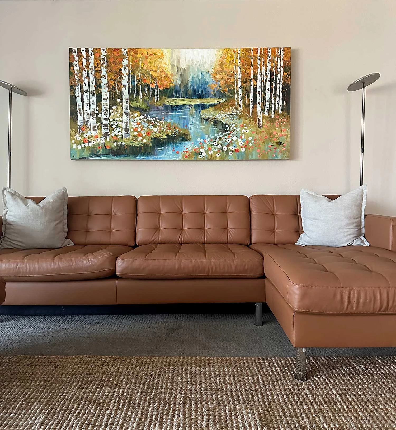 Living room with a tan leather sectional sofa, two white pillows, a colorful landscape painting of a forest with a river, and a brown woven rug.