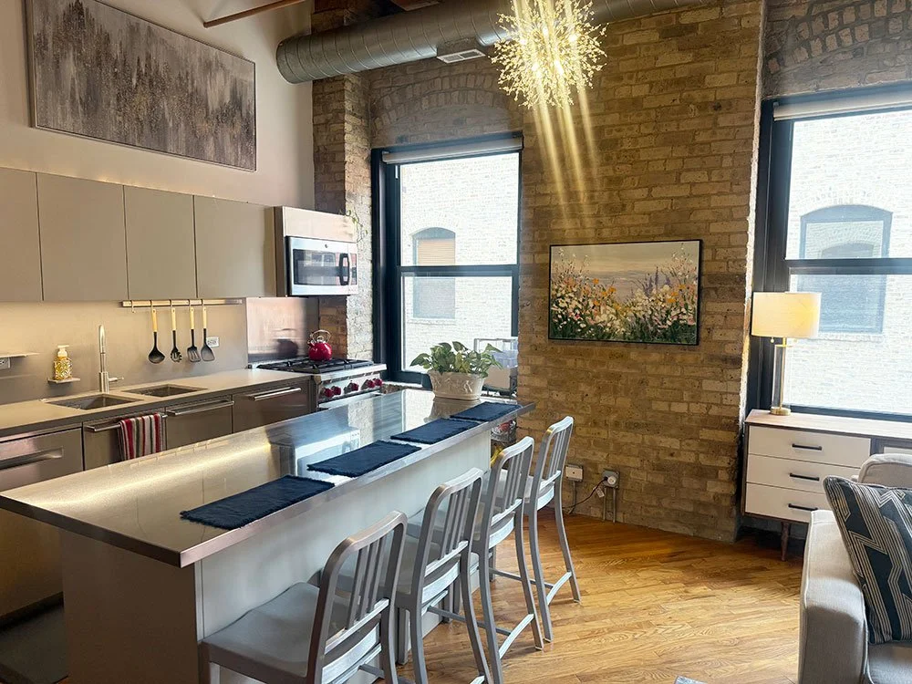 Modern kitchen with stainless steel appliances, a white island with four chairs, brick wall, large windows, abstract art, and a small white dresser with a lamp.