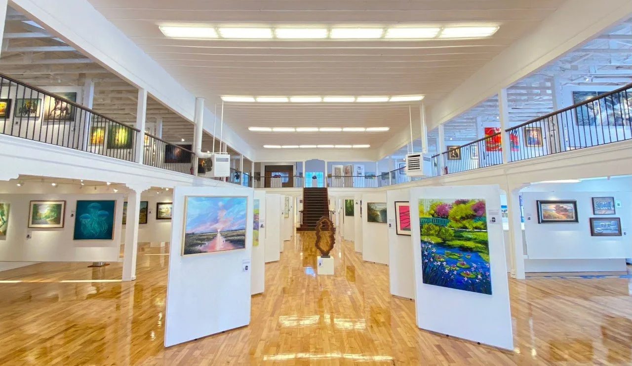 An art gallery with white partition walls displaying colorful paintings, a polished wooden floor, and a two-story open layout with artworks along the upper walkway.