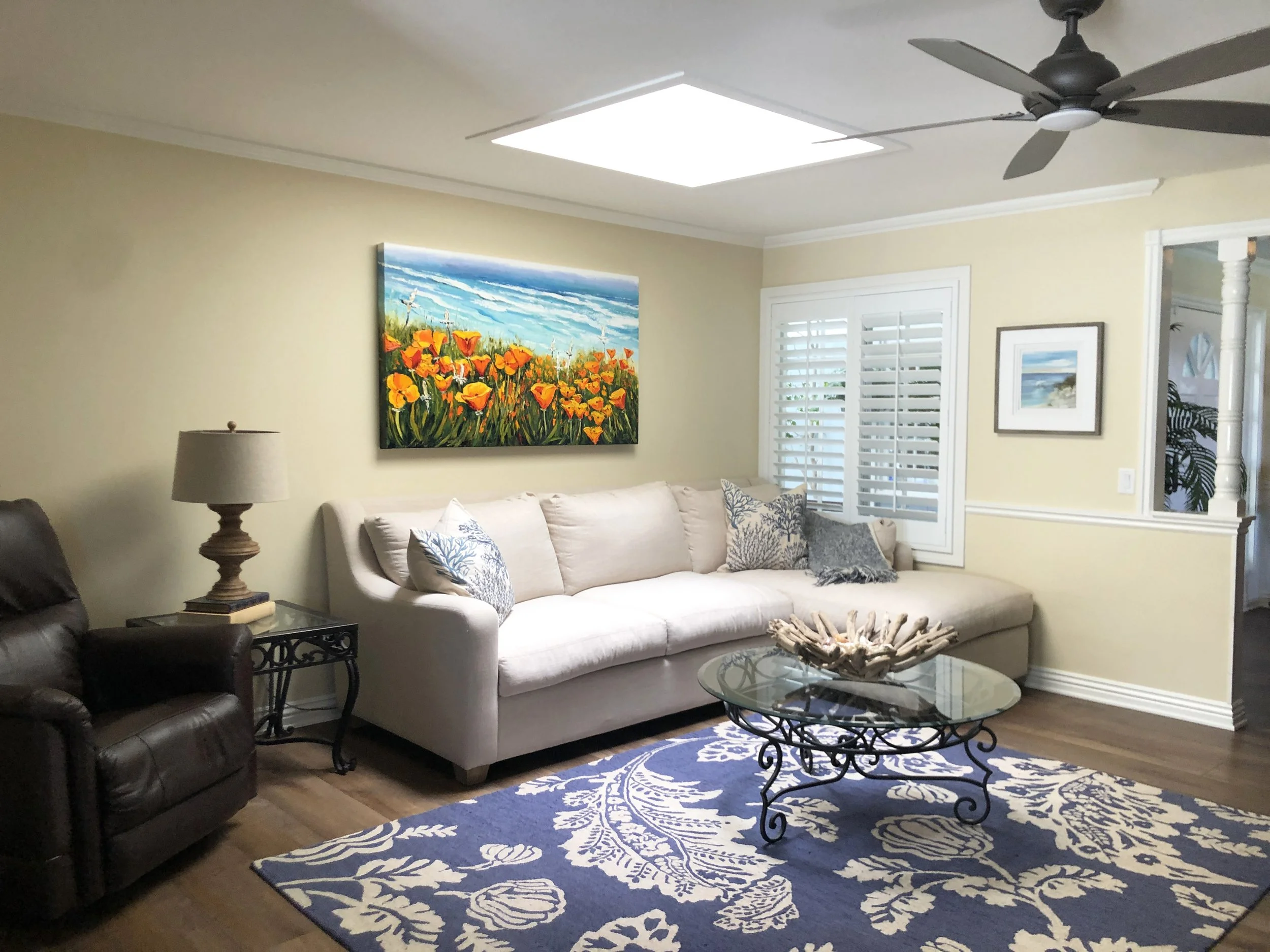 Living room with beige sofa, dark leather armchair, glass coffee table, floral rug, wall art of a flower field and ocean scene, ceiling fan, ceiling light, window with white blinds, table lamp, decorative items on side table, and a mirror.