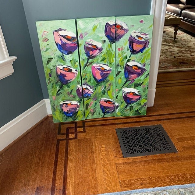 A four-panel abstract painting of colorful flowers in shades of pink, purple, and blue on a green background, leaning against a wall on a hardwood floor.