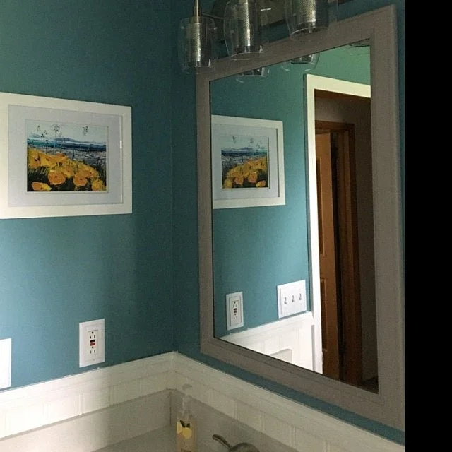 A bathroom mirror reflecting a teal wall with framed landscape art, electrical outlets, and a door.
