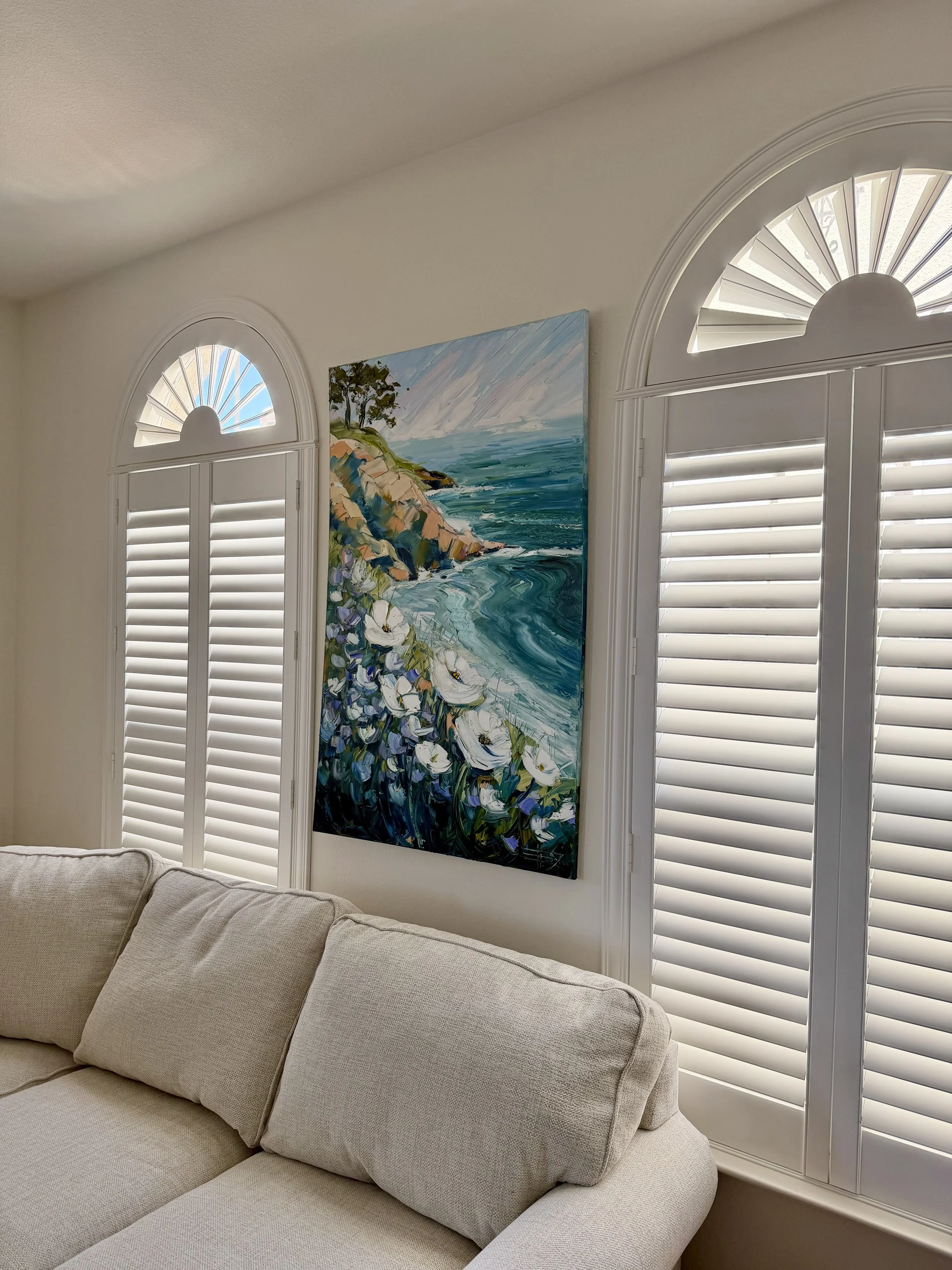 Living room corner with beige sofa, large window with white shutters, and a landscape painting of the coast with cliffs, trees, ocean, and sky.