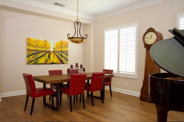 Dining room with a wooden table, six red chairs, a chandelier, a large wall clock, a three-pane window with shutters, a painting of a vineyard scene, and a black grand piano.