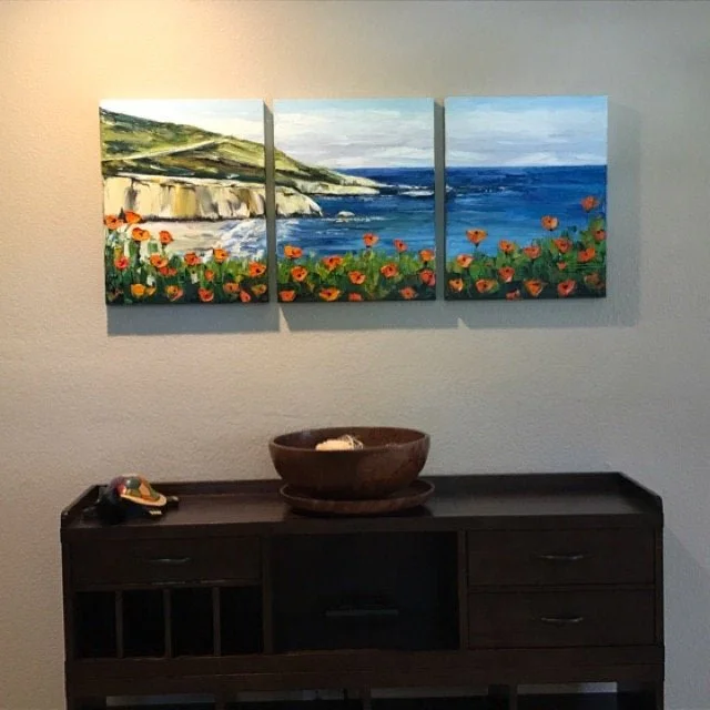 A three-panel landscape painting of a seaside scene with cliffs, green hills, and a blue ocean hangs on a light-colored wall above a dark wooden sideboard. The sideboard has a bowl, a remote, and some other small items on it.