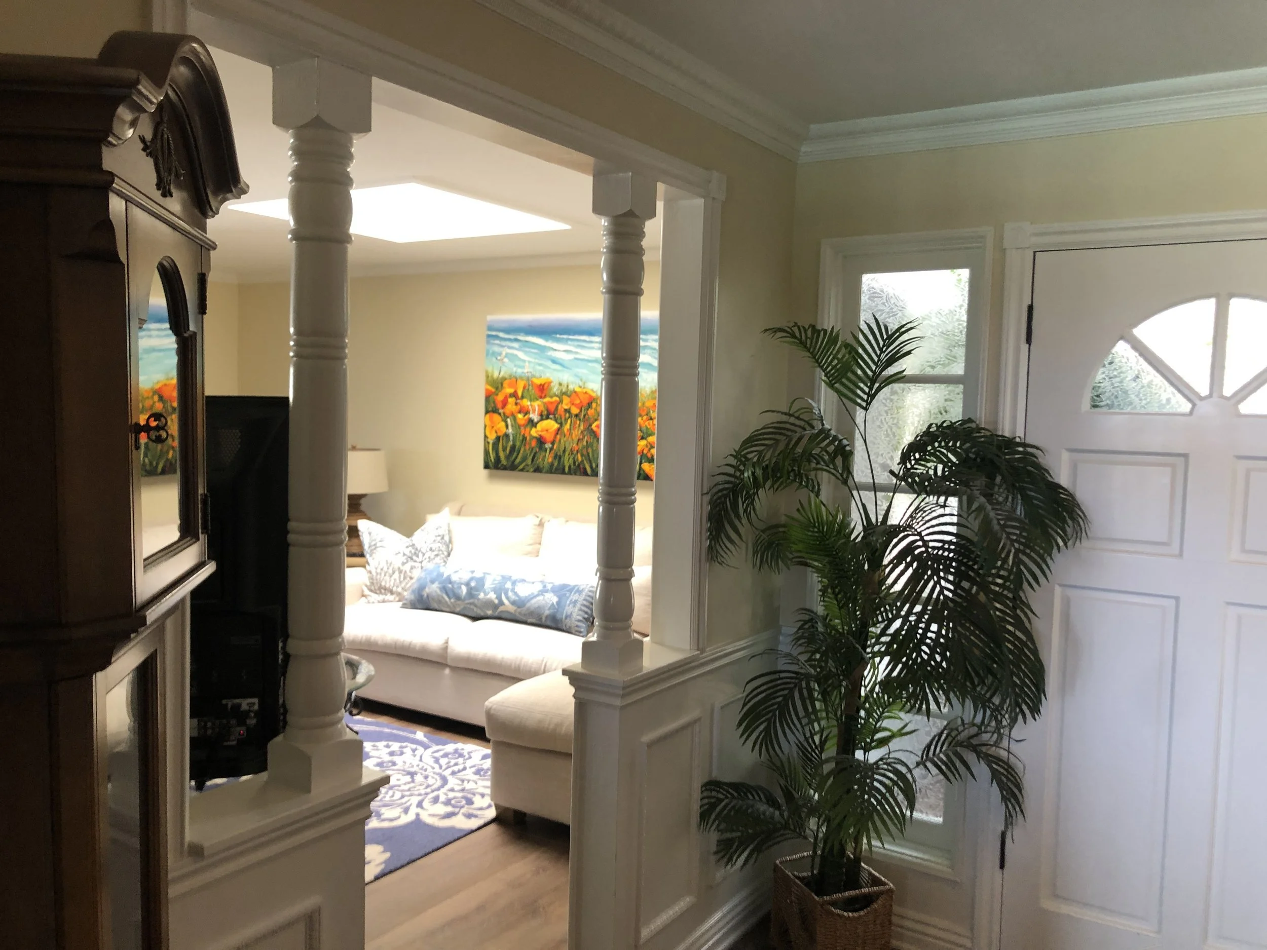 Living room with a white sofa, floral cushions, colorful painting of flowers and ocean, window with frosted glass, plant, and decorative columns.