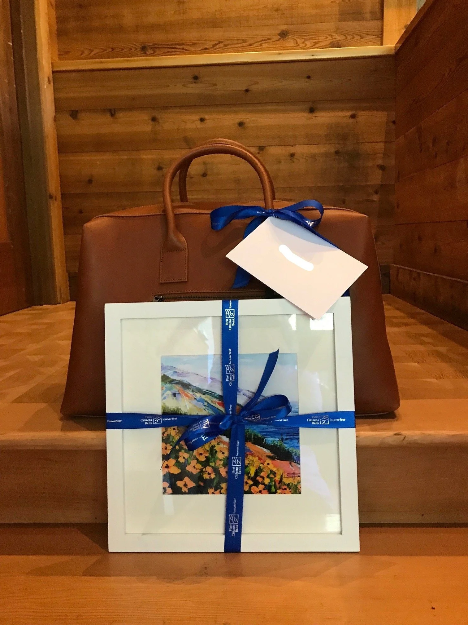 A boxed set of artwork with colorful landscape paintings, tied with a blue ribbon that has a logo, sitting on a wooden surface.