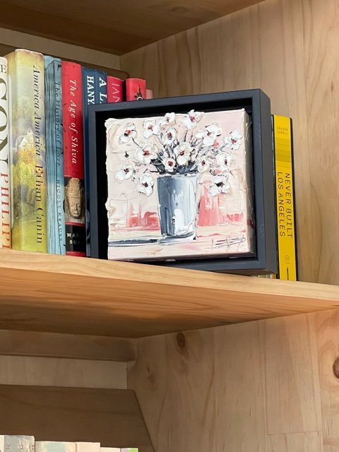 A framed painting of a white vase with white flowers on a shelf next to books in a wooden bookshelf.