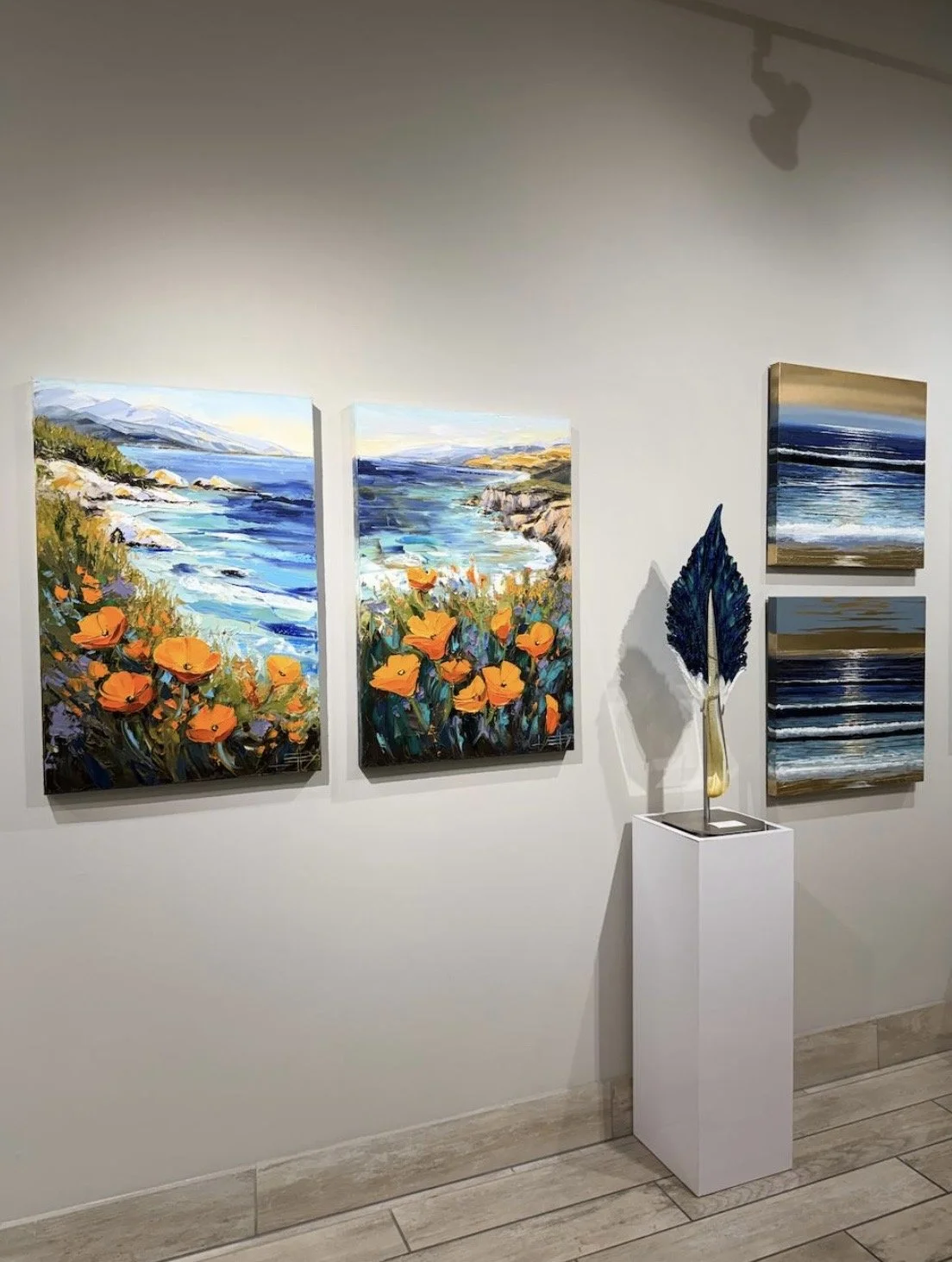 An art gallery wall with four landscape paintings of ocean scenes, flowers, and waves, along with a sculpture of a blue leaf on a pedestal.