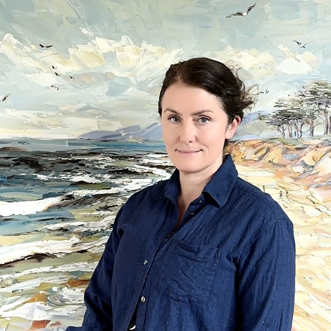A woman with dark hair in a bun wearing a dark blue denim shirt standing in front of an abstract landscape painting depicting a coastline with trees, rocks, and sky with clouds and birds.