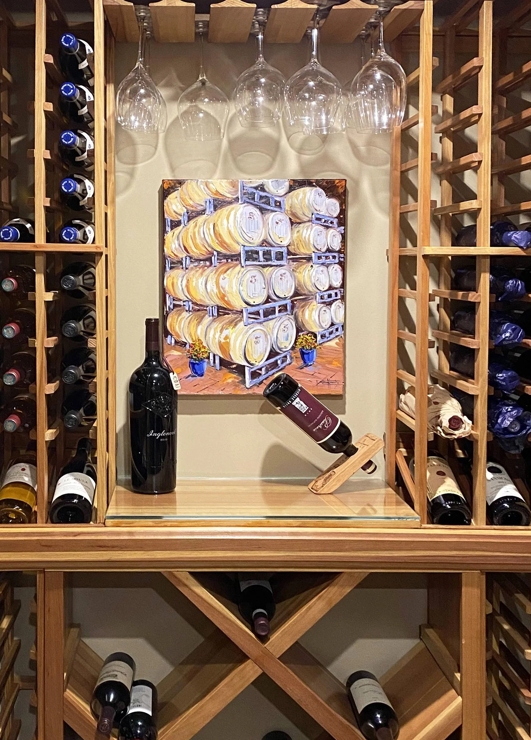 Display of wine bottles stored on wooden wine racks with empty wine glasses hanging upside down above, a painting of wine barrels on the wall, and a bottle of wine and a wine glass on the table.