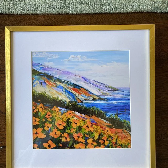 A framed painting of a coastal landscape featuring colorful flowers in the foreground, green hills, and blue ocean with mountains in the background under a partly cloudy sky.