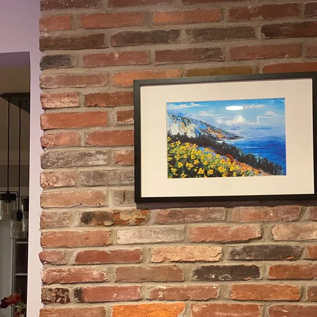 Painted landscape of cliffs, ocean, and yellow flowers in a frame hanging on a brick wall.
