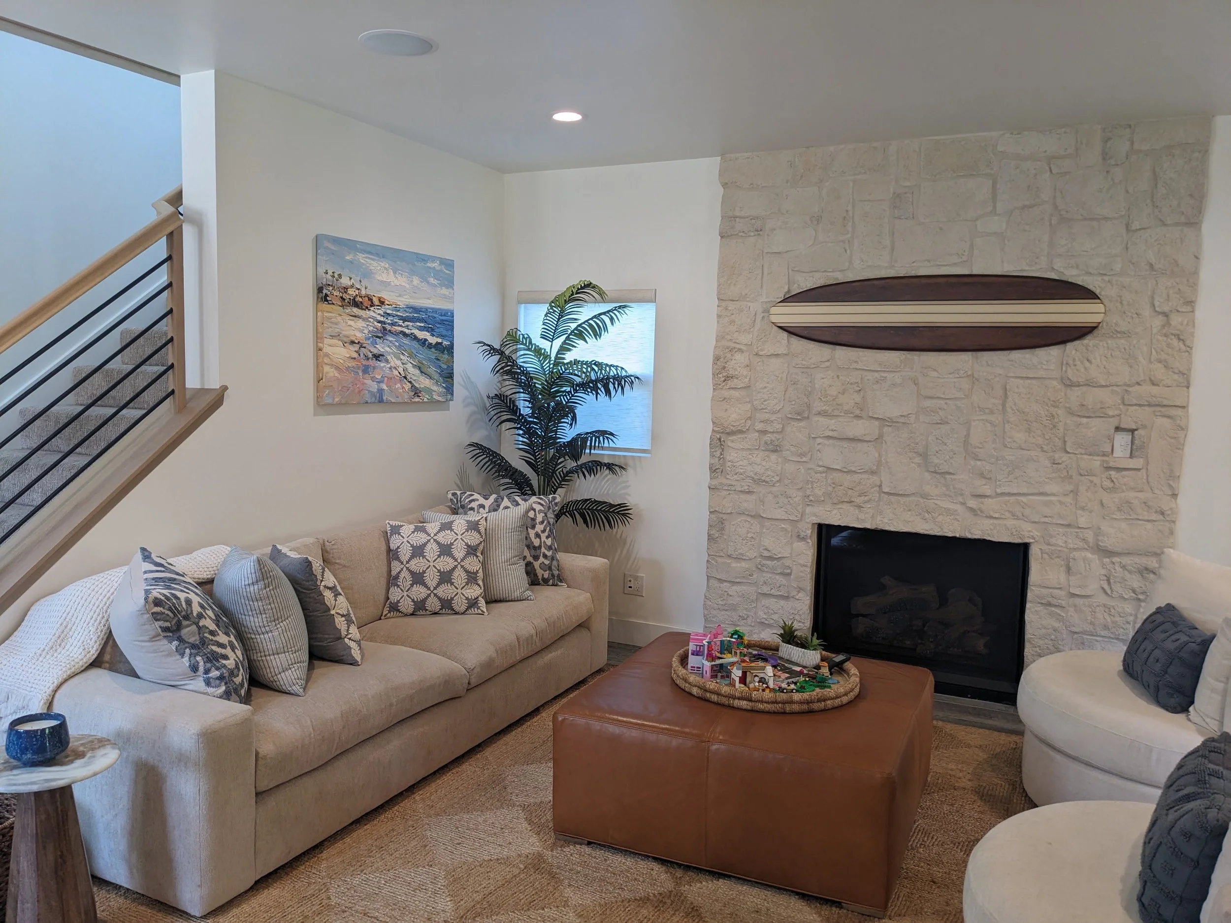A cozy living room with a beige sofa decorated with patterned pillows, a large tree, a stone fireplace with a wooden surfboard mounted above it, an artwork of a seascape, a window, and a children's play area with toys.