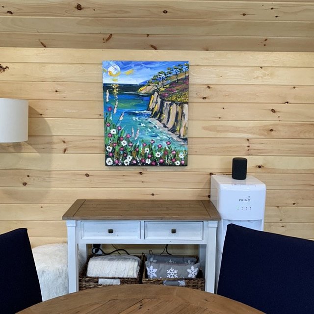 Interior of a room with wooden panel walls, featuring a colorful landscape painting of a river, trees, and flowers hung above a white table, with black chairs around a wooden dining table.
