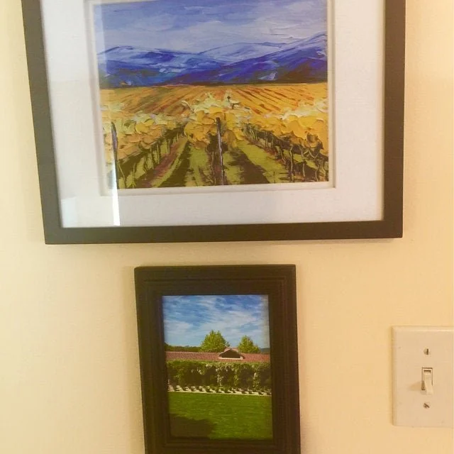 Two framed landscape paintings hanging on a beige wall, one above the other. The top painting depicts a vineyard with rolling hills and mountains in the background. The bottom painting features a manicured garden with a fountain, trees, and a cloudy 