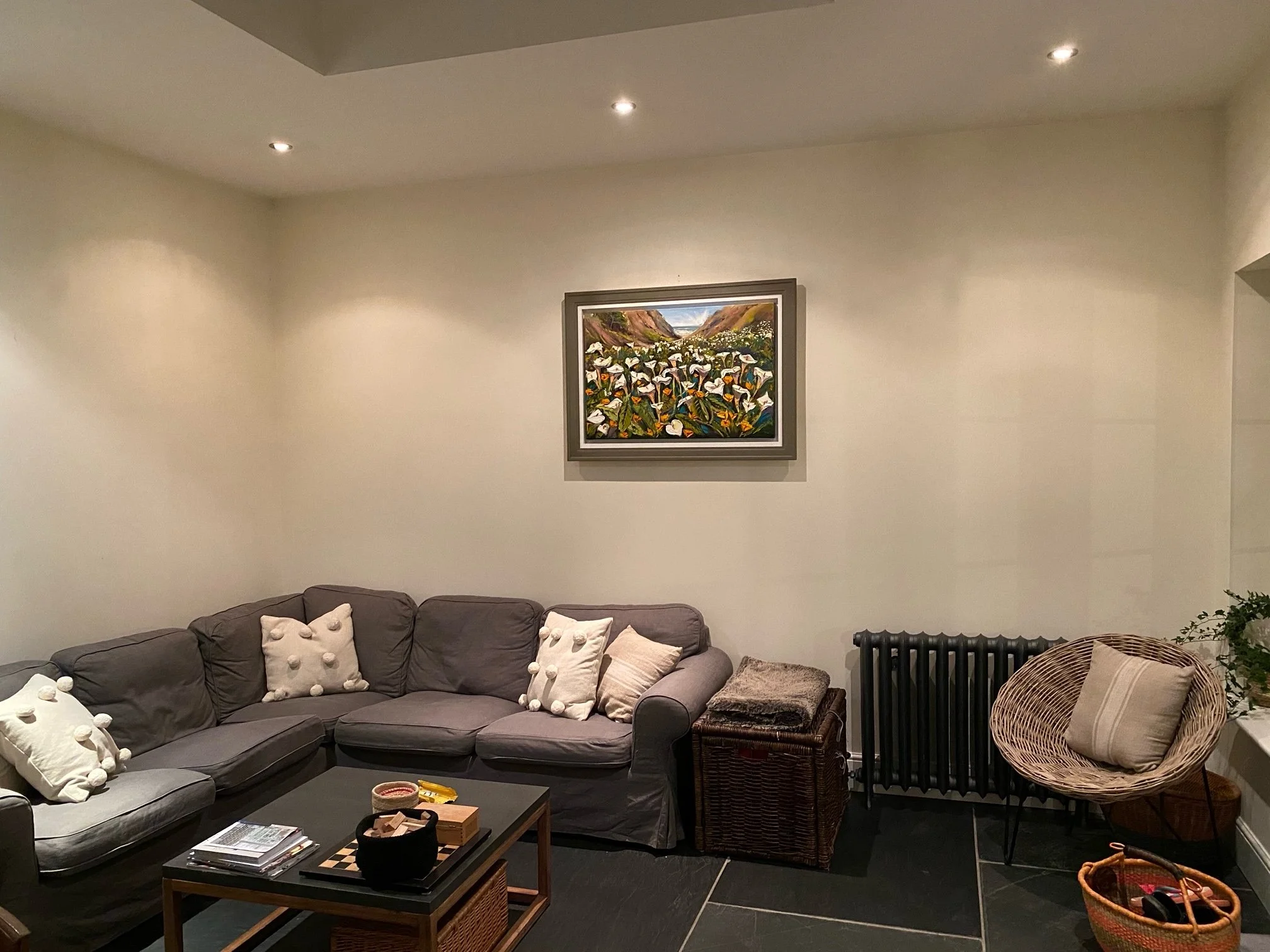 Living room with gray sectional sofa with multiple cream-colored pillows, a black coffee table with magazines, a wicker basket, and a framed landscape painting of flowers and mountains on the wall. There are woven chairs and a small plant to the side