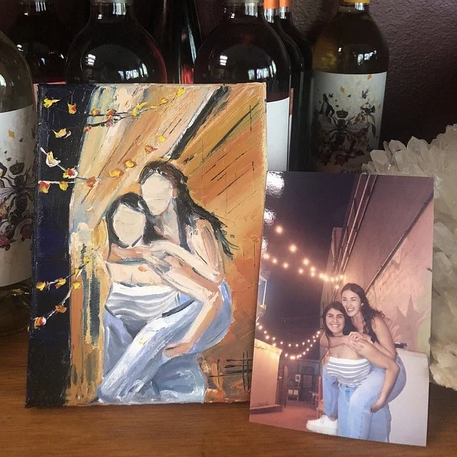 A hand-painted portrait of two women with blurred faces, sitting closely together, with a background of warm, colorful brushstrokes and a string of lights. Beside the painting is a photograph of two women smiling and hugging outdoors at night, with s