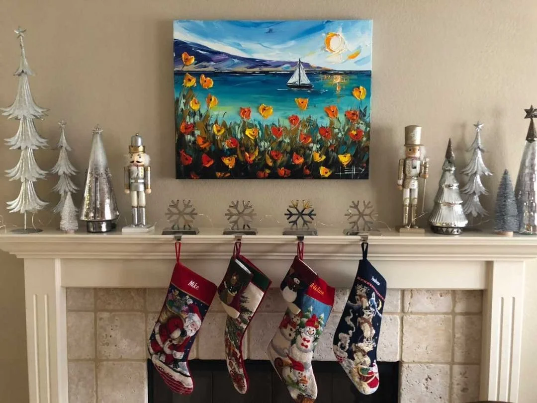 Christmas mantel decorated with silver and white trees, nutcracker figures, snowflake ornaments, and hanging stockings featuring festive Christmas designs, with a colorful landscape painting of a sailboat on water above.