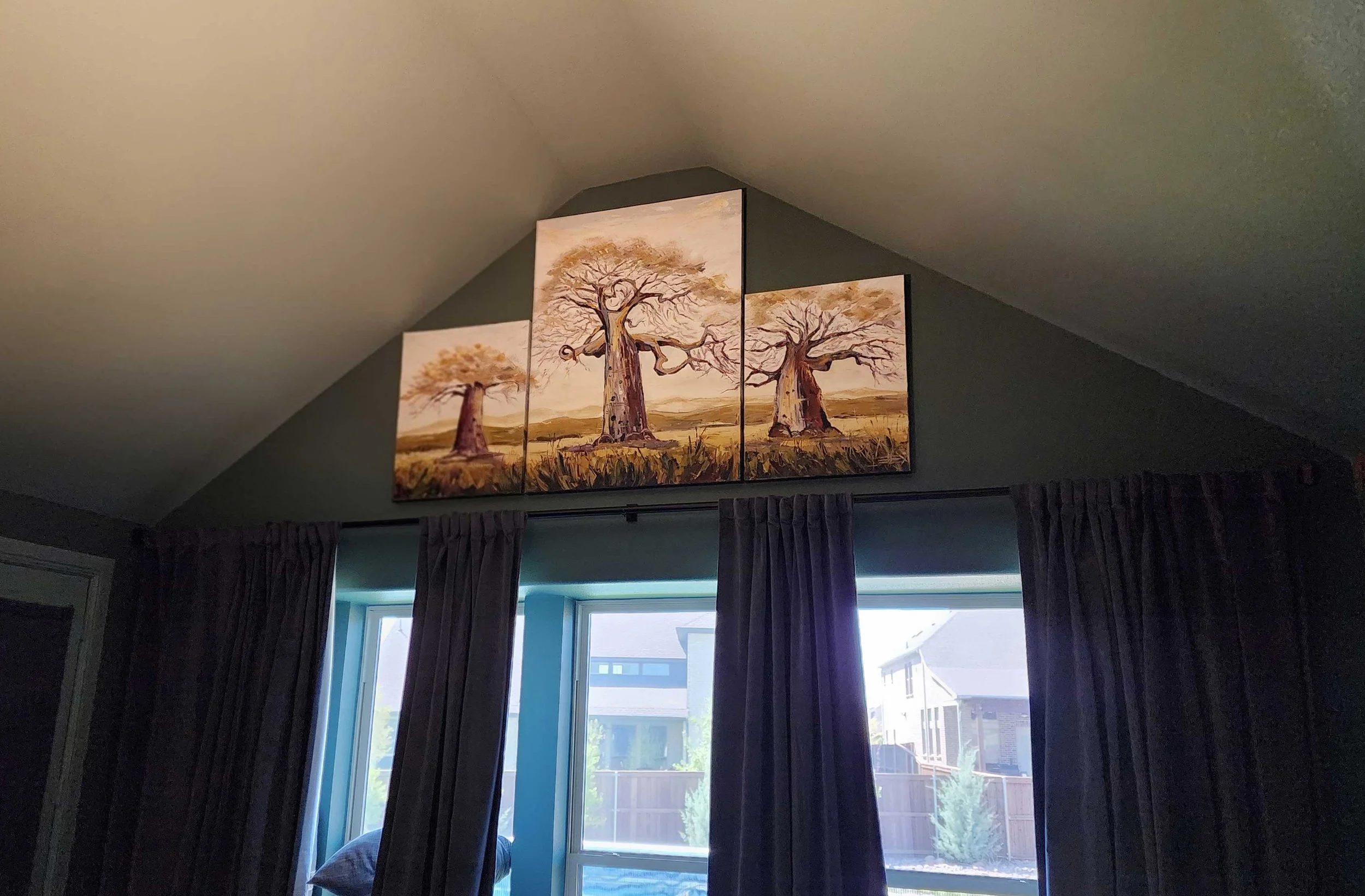 Three-panel painting of leafless trees in a landscape, hung on a sloped ceiling in front of a window with curtains, with parts of the window and houses outside visible.
