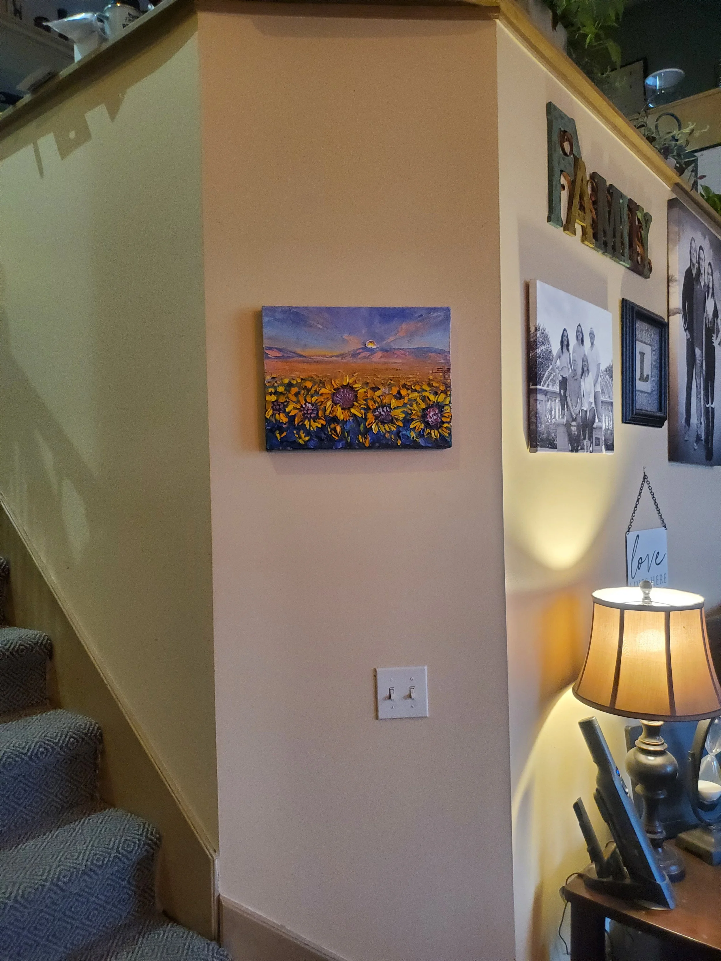 A decorated wall with a framed sunflowers painting, black-and-white family photo, and colorful 'FAMILY' sign. A lamp with a beige lampshade is on a small table, and an exercise bike is nearby.