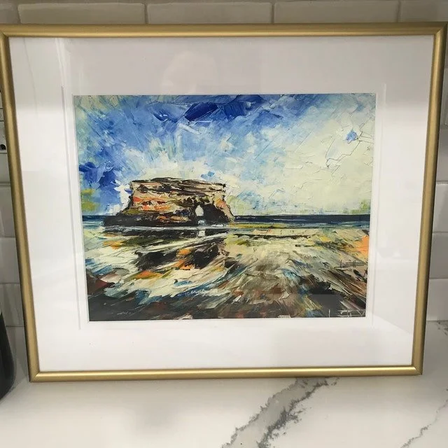 Framed landscape painting of a coastal scene with a rocky island, sea, and sky painted in an impressionistic style.